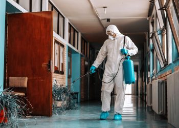Full length of worker in sterile uniform, with face mask holding sprayer with disinfectant and spraying school hallway.