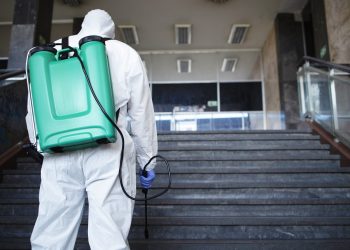 Unrecognizable person in white chemical protection suit with reservoir disinfecting public corridor to stop spreading highly contagious corona virus.
